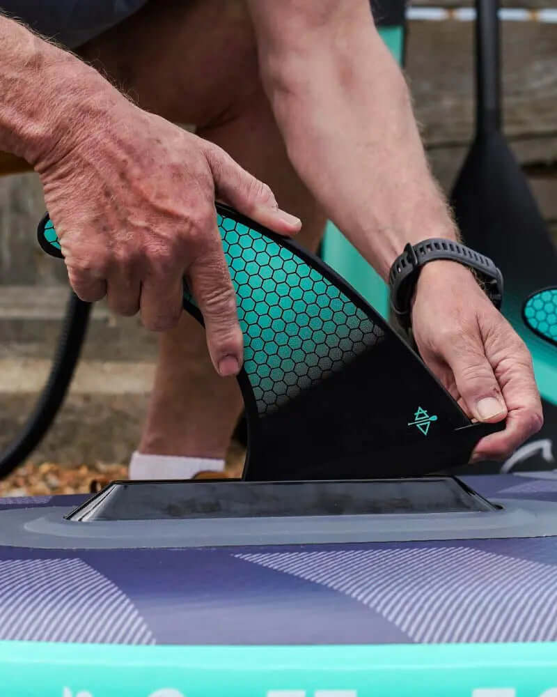 Person befestigt eine schwarze Flosse an einem aufblasbaren Paddleboard mit strukturierter Oberfläche