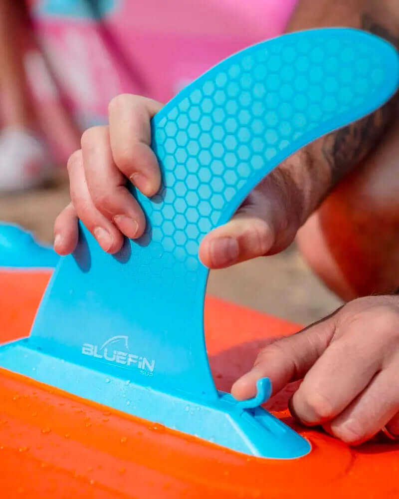 Person attaching a blue fin to an orange inflatable paddleboard with water droplets.