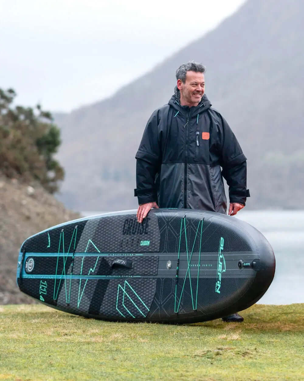 Man in black jacket standing on grass with a Bluefin inflatable paddleboard, mountains in background