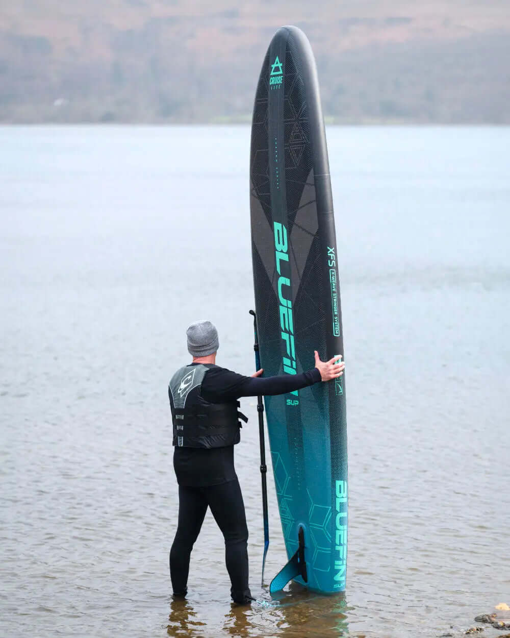 Person in wetsuit holding a Bluefin inflatable paddleboard in shallow water, wearing a life vest and beanie.