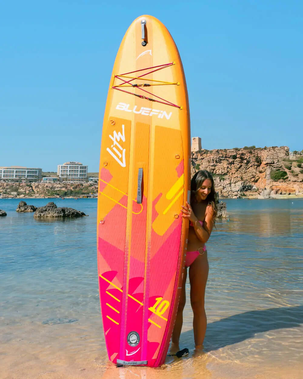 Inflatable paddleboard with vibrant orange and pink design, woman holding it in shallow water