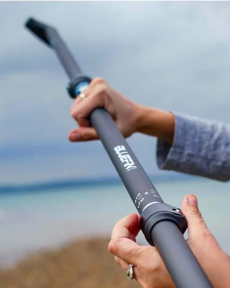 Person, die ein schwarzes verstellbares Paddel mit strukturiertem Griff und gebogenem Ende vor einem bewölkten Himmel hält.