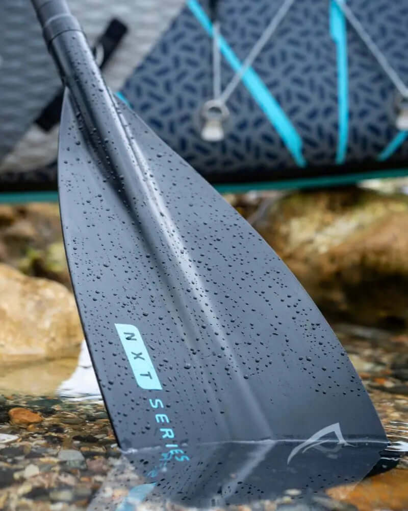 Nahaufnahme einer schwarzen Paddelblatt mit Wassertropfen, beschriftet NXT-Serie, neben einem Paddleboard.