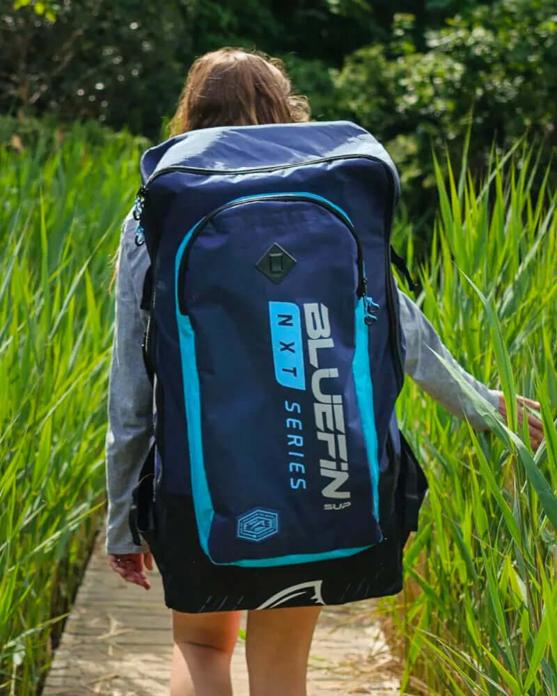 Person walking on a path with tall grass, carrying a blue and black Bluefin SUP backpack.