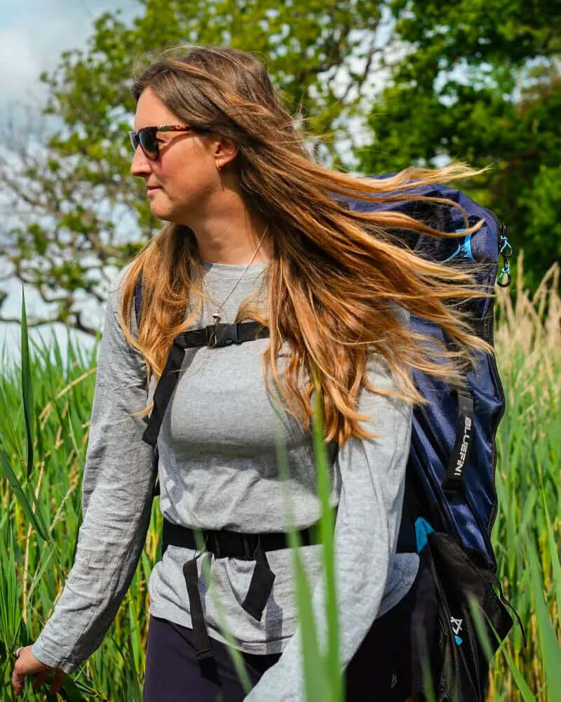 Woman with long hair wearing sunglasses and a gray shirt, carrying a backpack through tall grass.