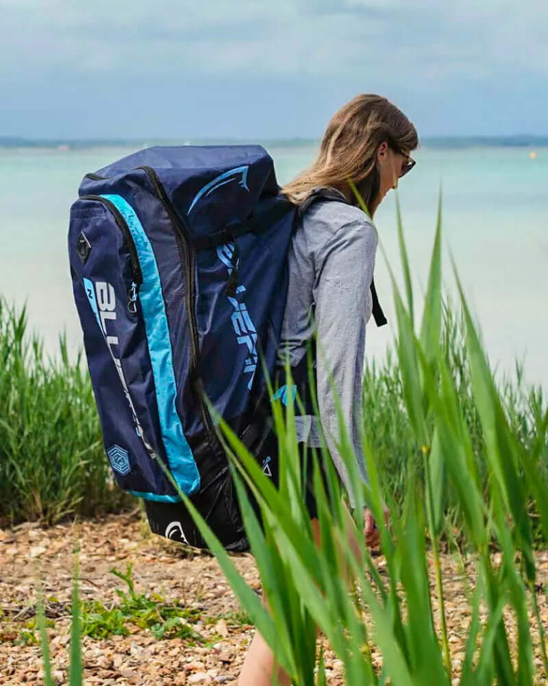 Person walking by the water carrying a large blue and black backpack with a logo