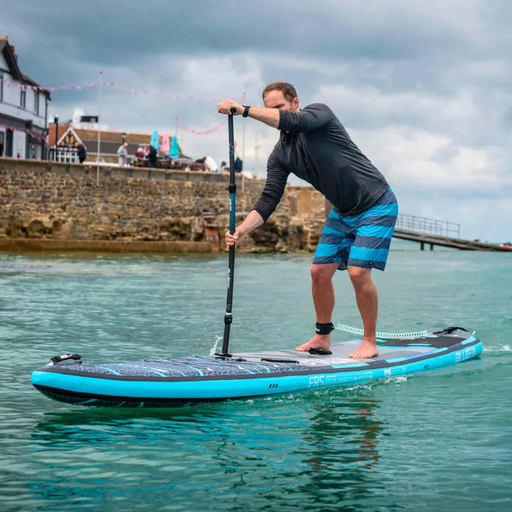 Mann paddelt auf einem blauen aufblasbaren Paddleboard im ruhigen Wasser, trägt ein schwarzes Shirt und gestreifte Shorts.