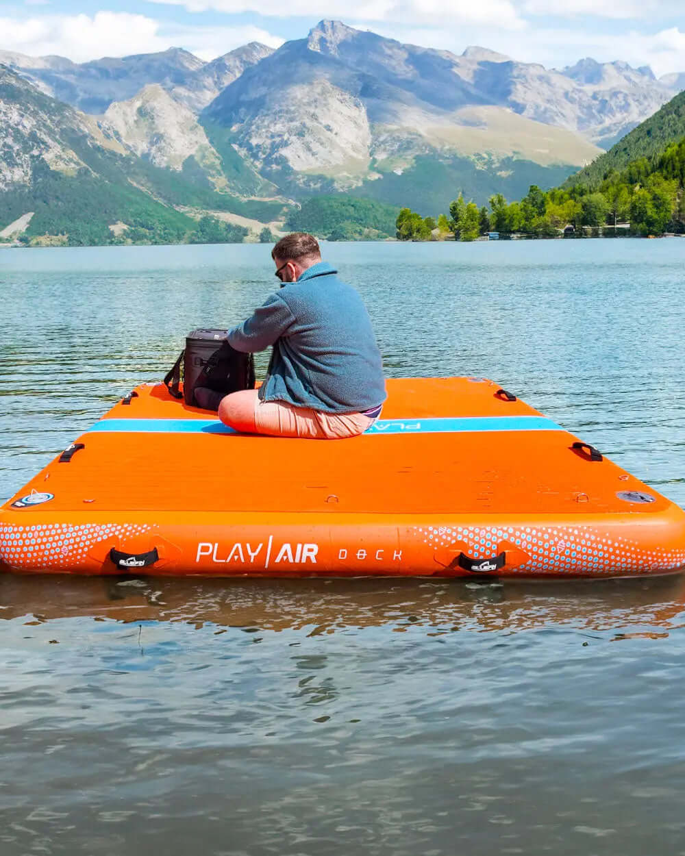 Person sitting on an orange inflatable dock in a lake with mountains in the background