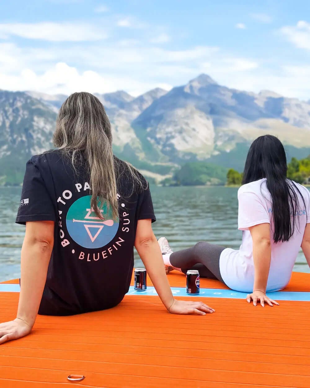 Two women sitting on an orange paddleboard by a lake, mountains in the background, enjoying drinks.
