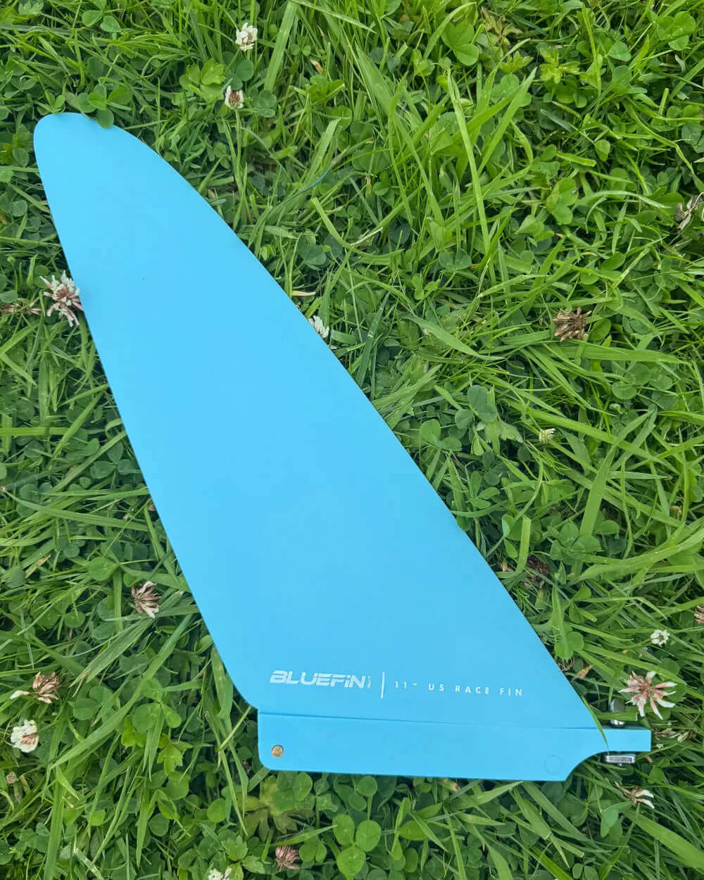 Bluefin 11" US race fin in bright blue color resting on green grass with small white flowers.