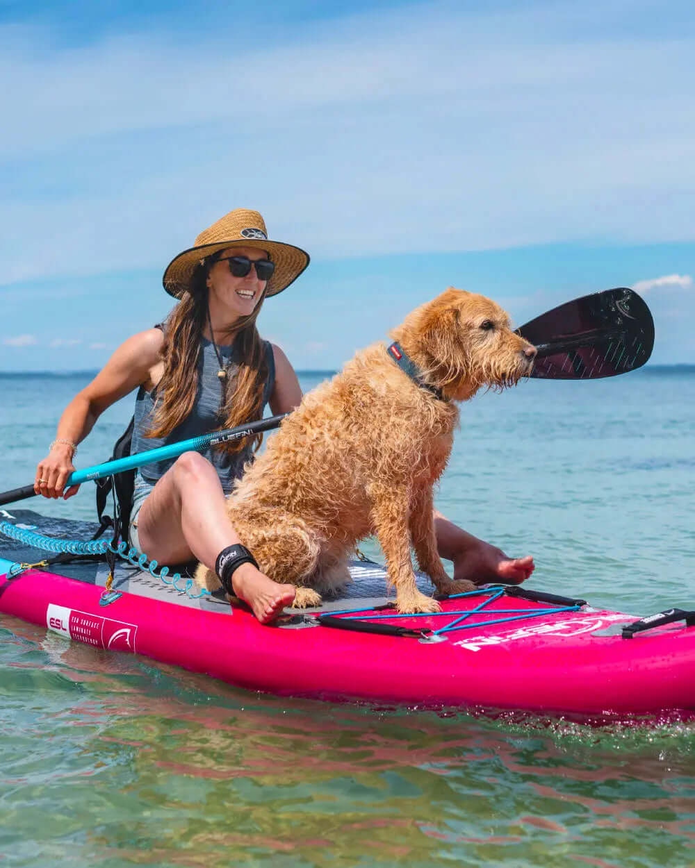 Woman in a hat paddling on a pink inflatable paddleboard with a dog sitting beside her in clear water.