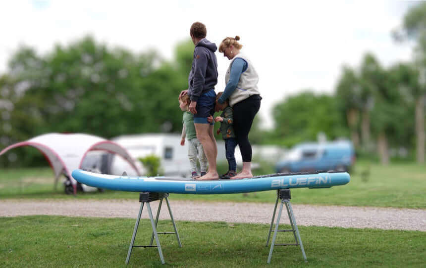 Family standing on a Bluefin paddleboard on a support frame in a grassy area.