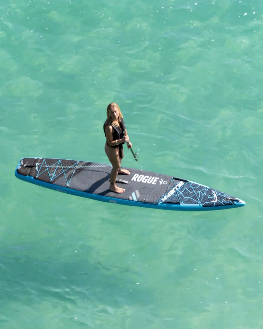 Person standing on a black and blue paddleboard in clear turquoise water holding a paddle