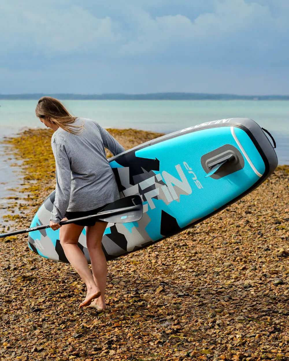 Woman carrying a blue and gray inflatable paddleboard along a rocky shore