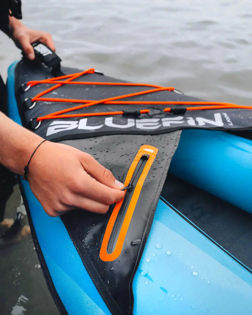 Hand securing an orange zipper on a black paddleboard deck with blue sides and water droplets.