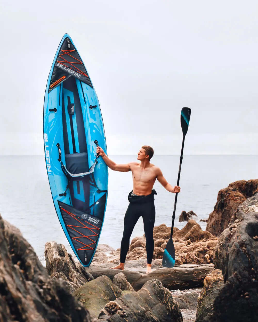 Man holding a blue inflatable paddleboard on rocky shore, wearing black wetsuit and holding a paddle
