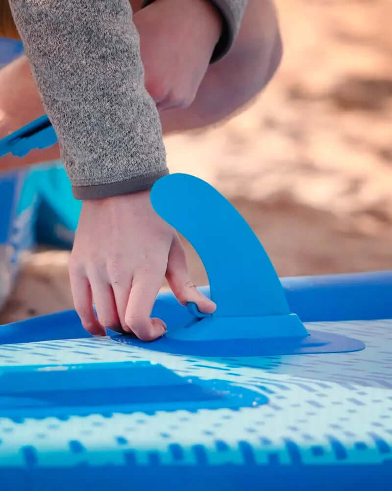 Person attaching a blue fin to an inflatable paddleboard on a sandy surface