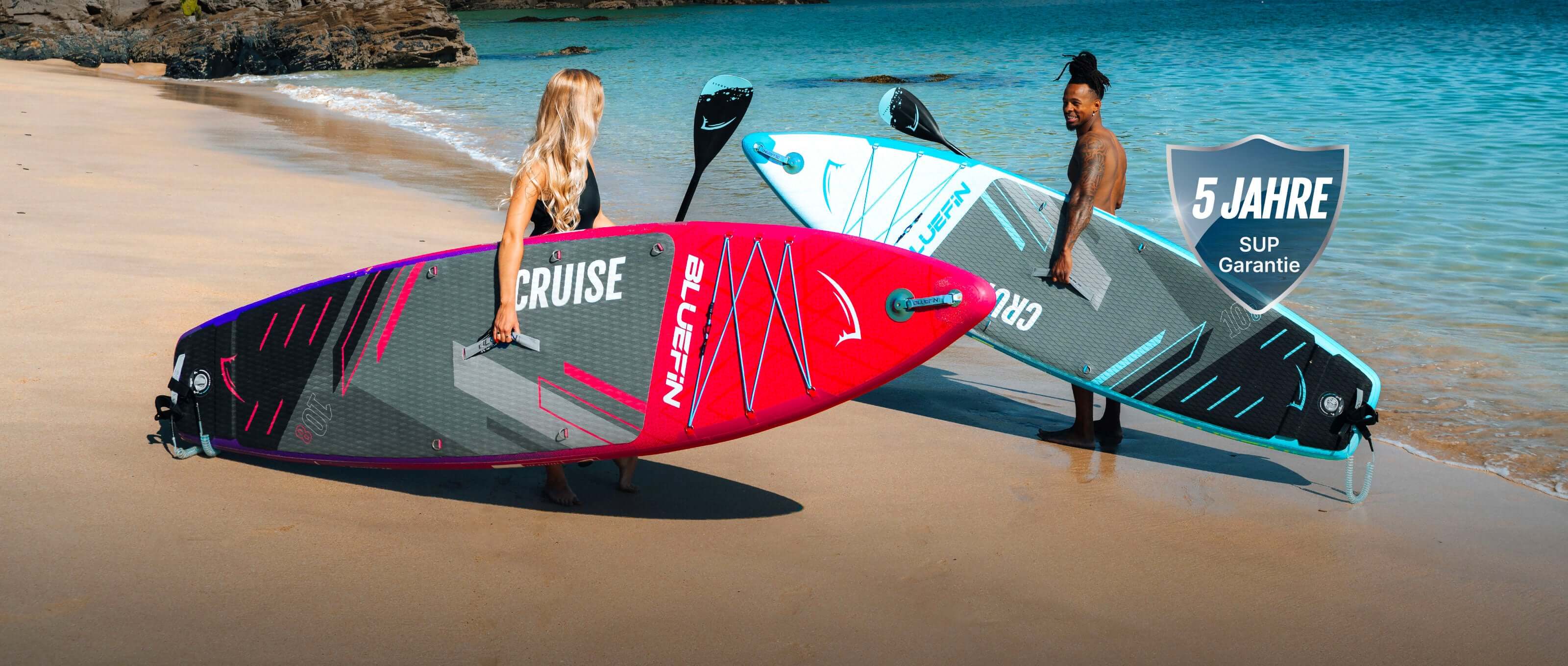 Two inflatable paddleboards on a beach, one pink and one blue, with paddlers standing beside them.