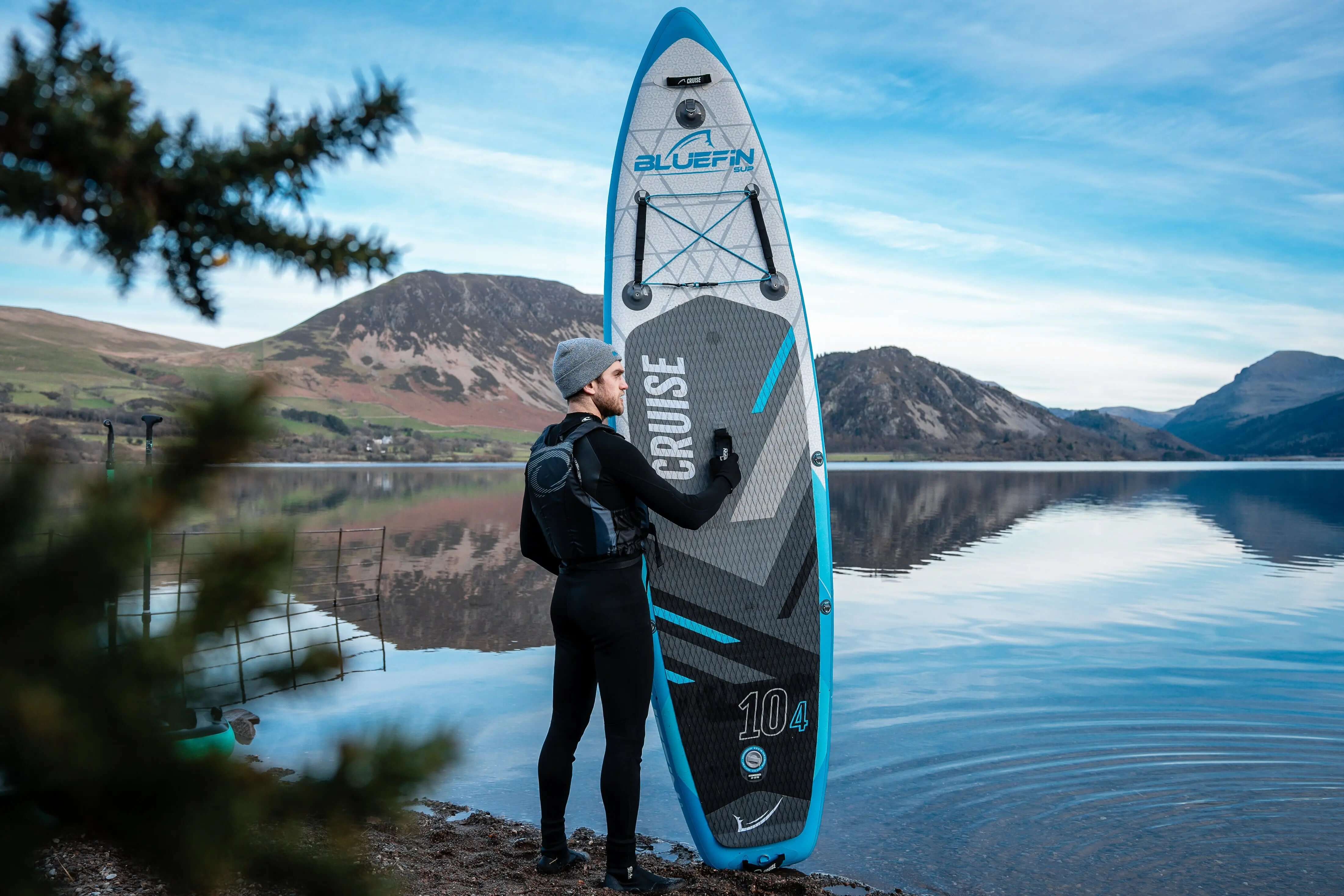 Person im Neoprenanzug hält ein Bluefin SUP Paddleboard am See mit Bergen im Hintergrund.