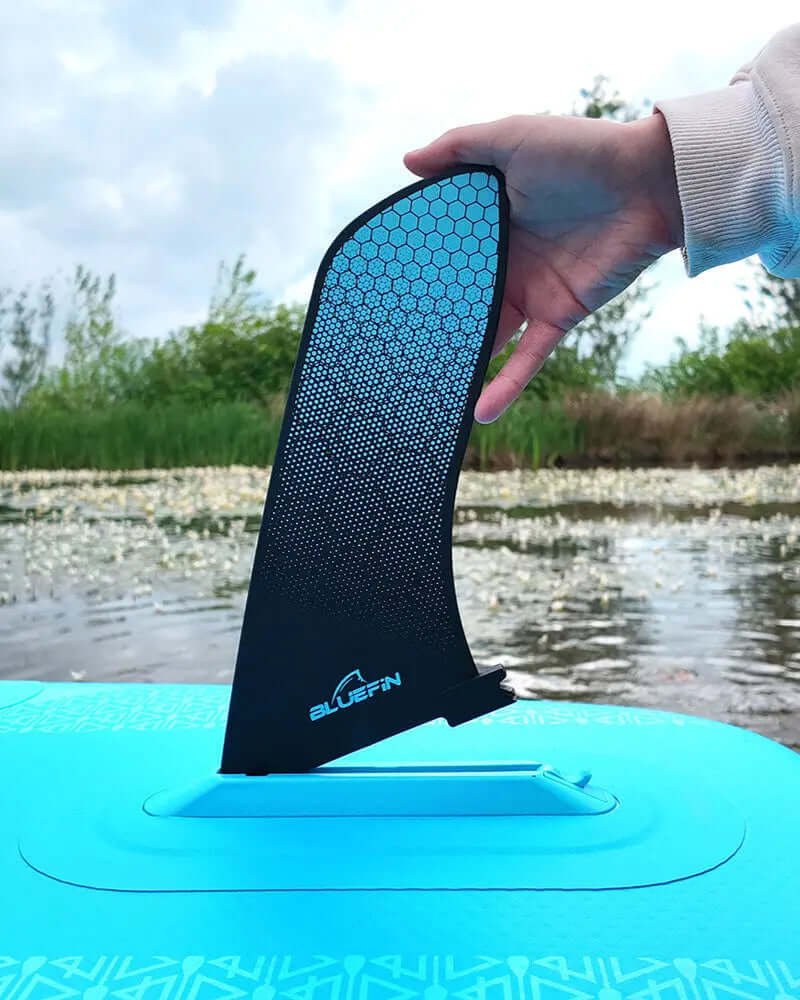 Hand holding a black fin with a blue honeycomb pattern above a blue inflatable paddleboard on water.