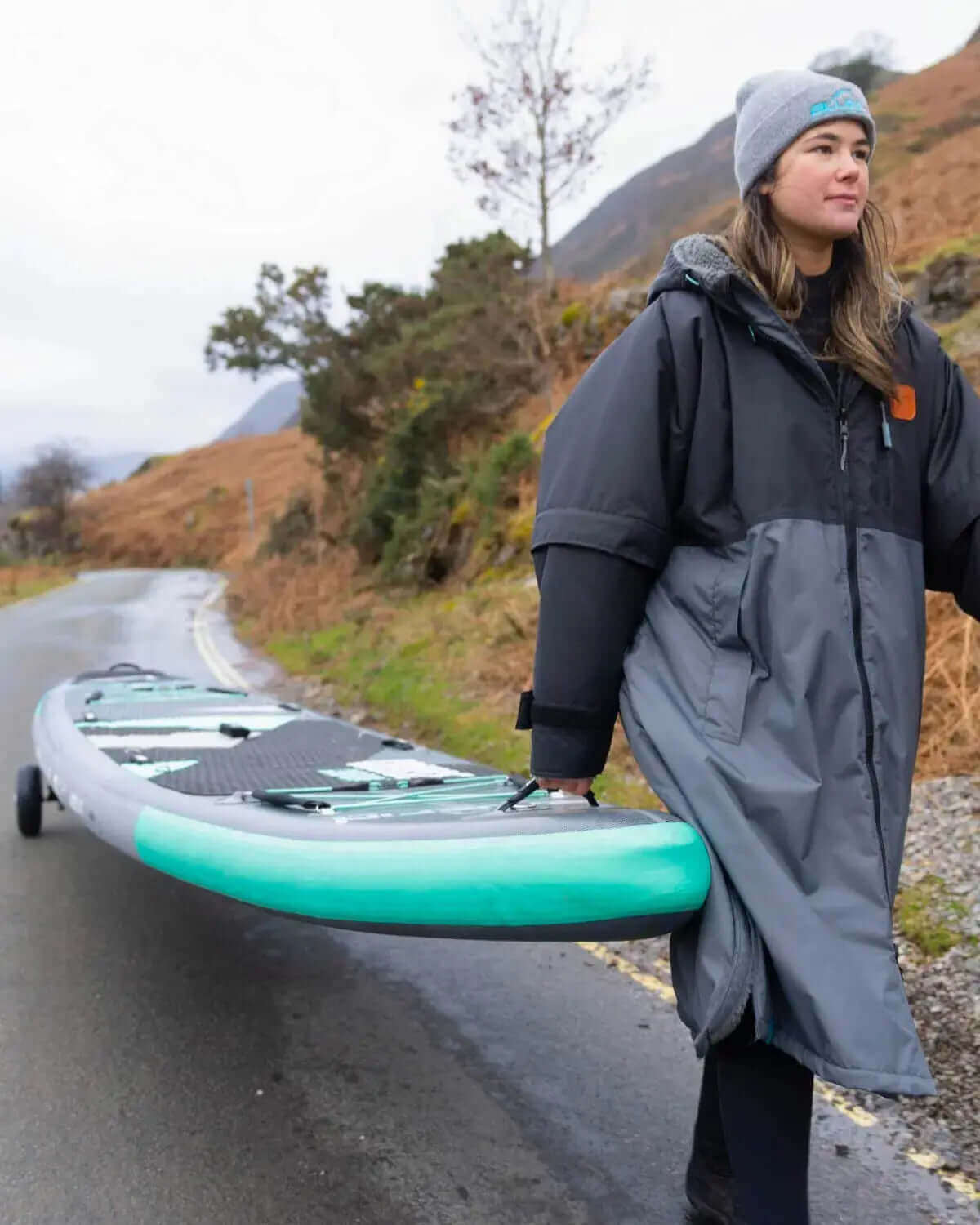 Person in a gray coat and beanie carrying a teal inflatable paddleboard along a road