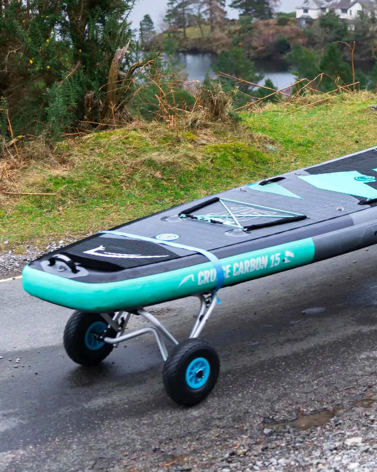 Inflatable paddleboard with wheels, textured deck, and blue accents on a path near a lake.