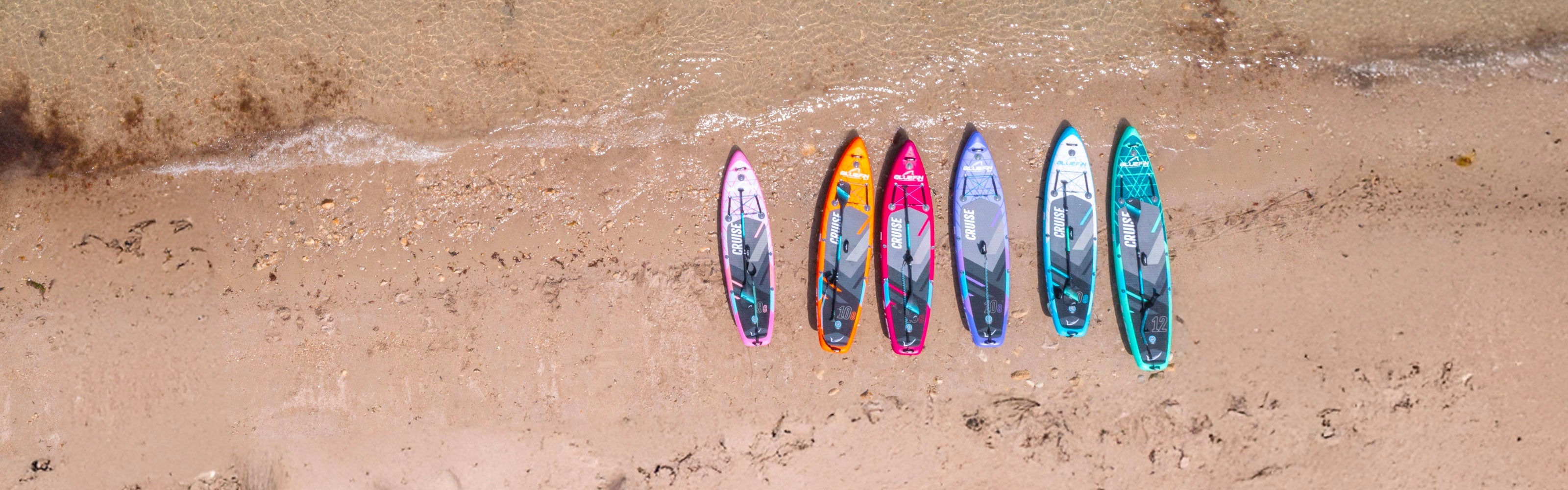 Six colorful inflatable paddleboards lined up on a sandy beach near the water's edge