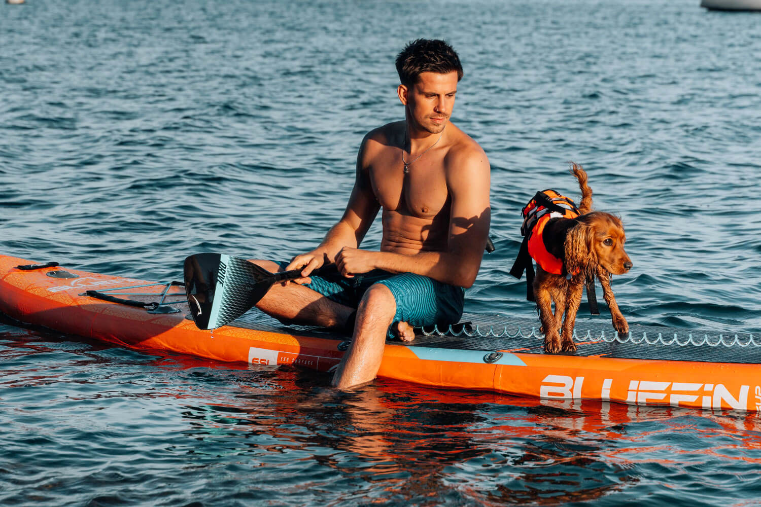 Man on orange paddleboard with a dog in a life jacket, both on calm water