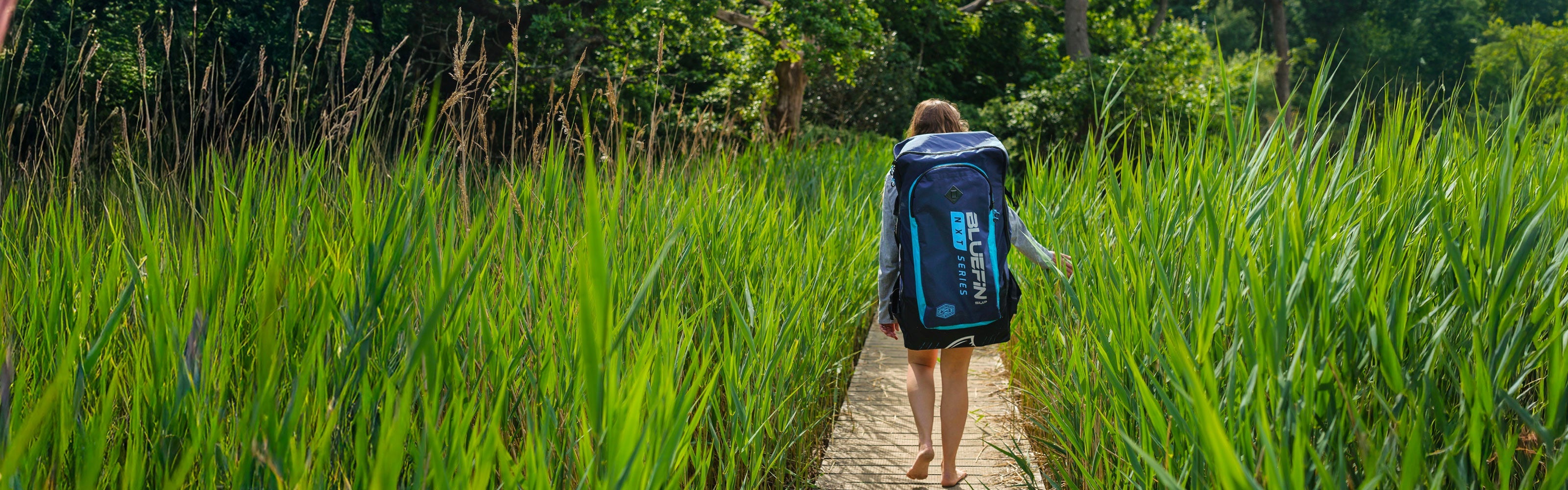 Person walking barefoot along a wooden boardwalk surrounded by tall green reeds, carrying a Bluefin inflatable paddleboard backpack, heading towards an outdoor adventure.