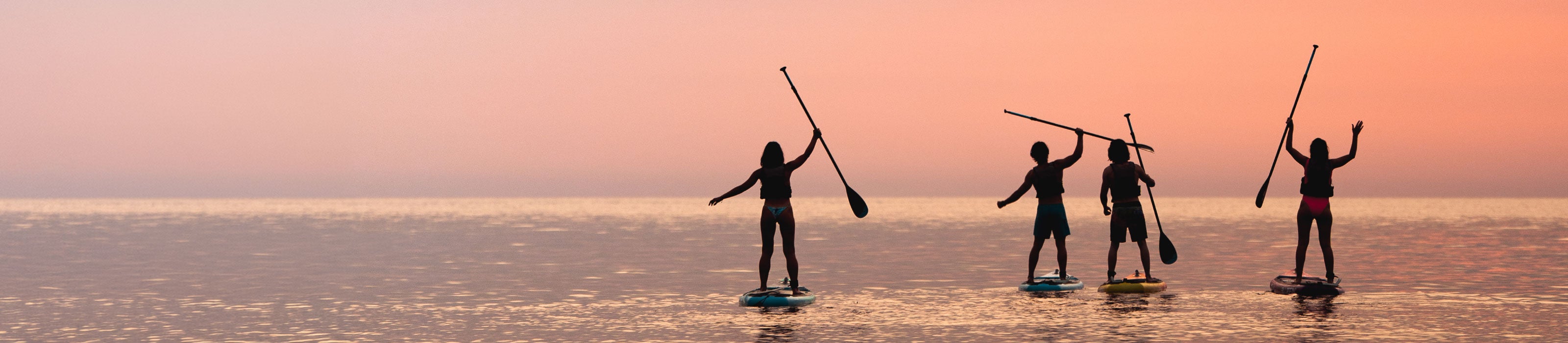 Four people paddleboarding at sunset, silhouetted against a pink sky on calm water.