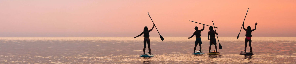 Vier Personen beim Stand-up-Paddling bei Sonnenuntergang, als Silhouetten vor rosa Himmel auf ruhigem Wasser