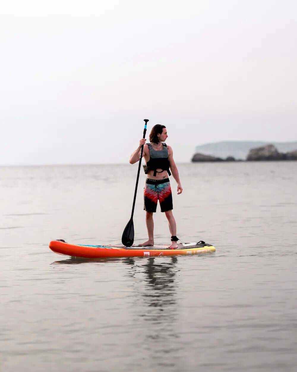 Person on an inflatable paddleboard in the water, holding a paddle, wearing a life jacket and swim shorts.