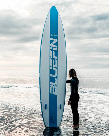 Person in wetsuit standing beside a blue and white Bluefin SUP paddleboard on the shore