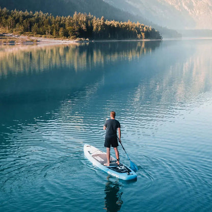 Person paddling on a blue stand-up paddleboard in calm water with forested mountains in the background