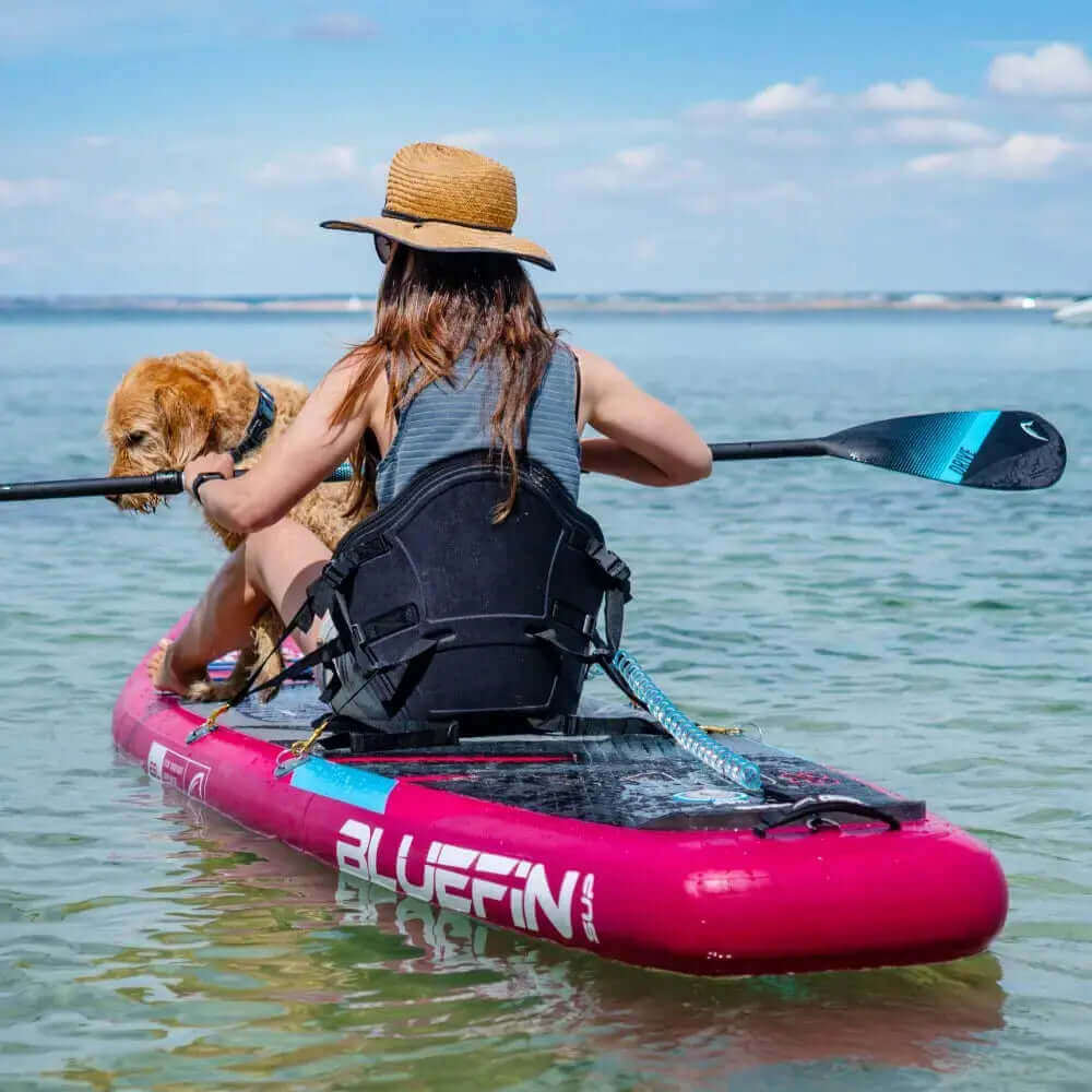Person, die auf einem rosa aufblasbaren Paddleboard mit einem Hund, der einen Strohhut trägt, in klarem Wasser paddelt.