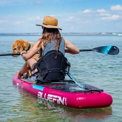 Person, die auf einem rosa aufblasbaren Paddleboard mit einem Hund, der einen Strohhut trägt, in klarem Wasser paddelt.