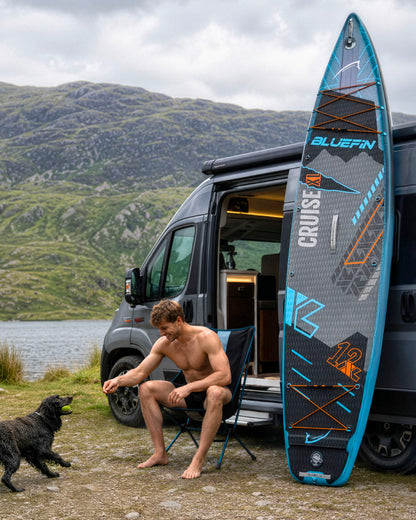 Man sitting in a chair by a van, playing with a dog near a Bluefin inflatable paddleboard.