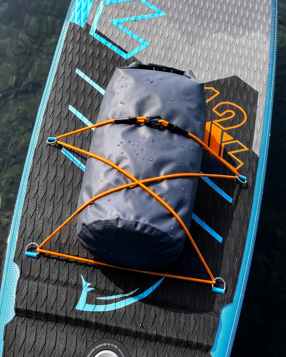 Paddleboard with a dark waterproof bag secured by orange bungee cords on a textured surface.