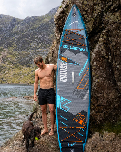 Man standing next to a Bluefin Cruise XL paddleboard on rocky shore, dog nearby, mountains in background.