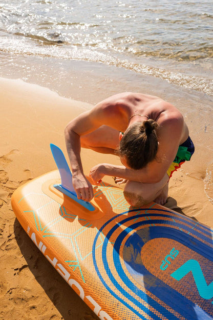 Person adjusting the blue fin on an orange inflatable paddleboard on sandy beach