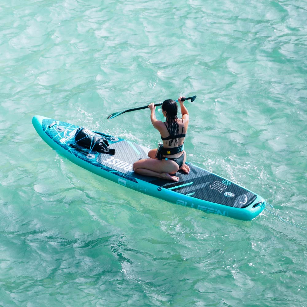 Person paddling on a blue inflatable paddleboard in clear water, wearing a black bikini.