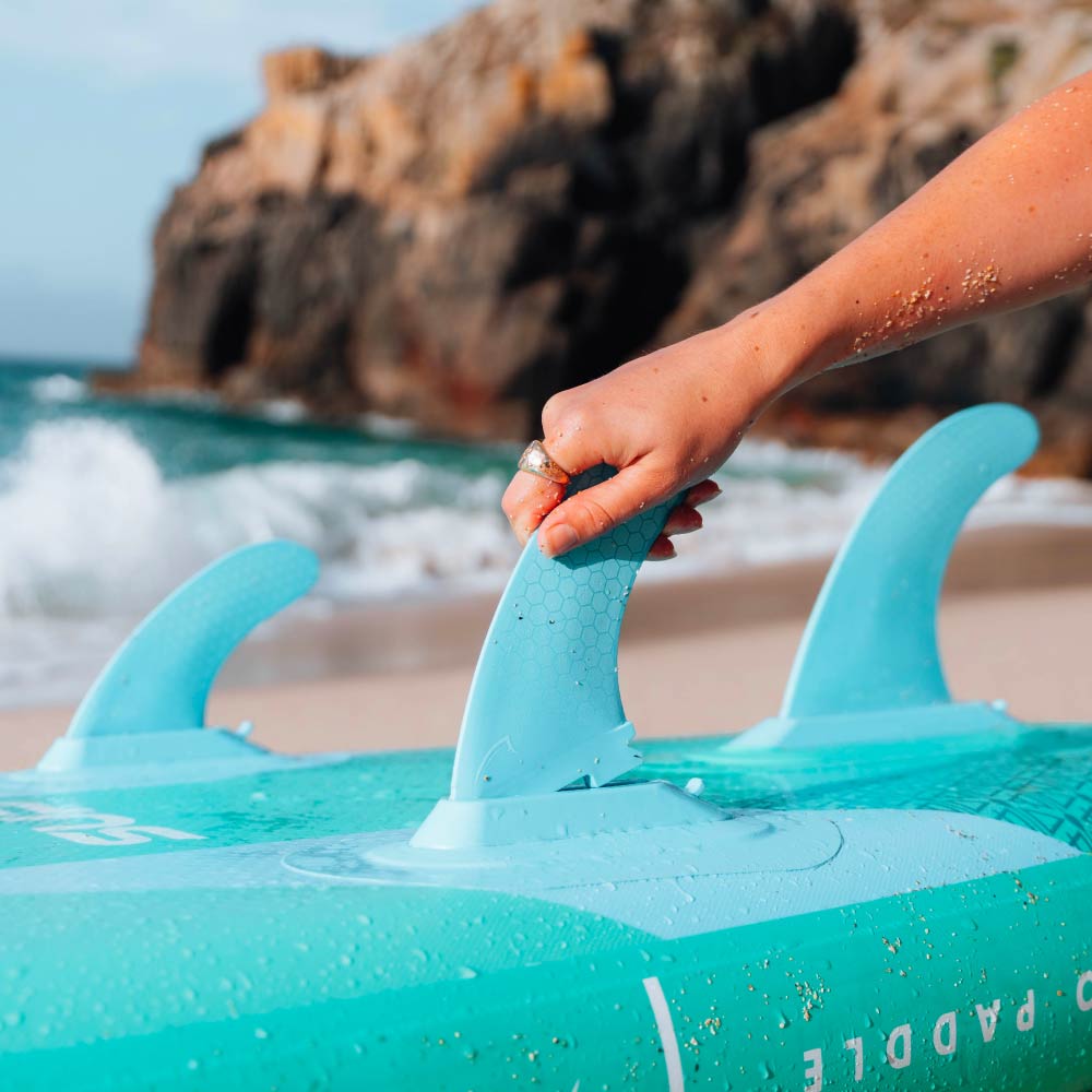 Hand adjusting the center fin on a turquoise inflatable paddleboard at the beach