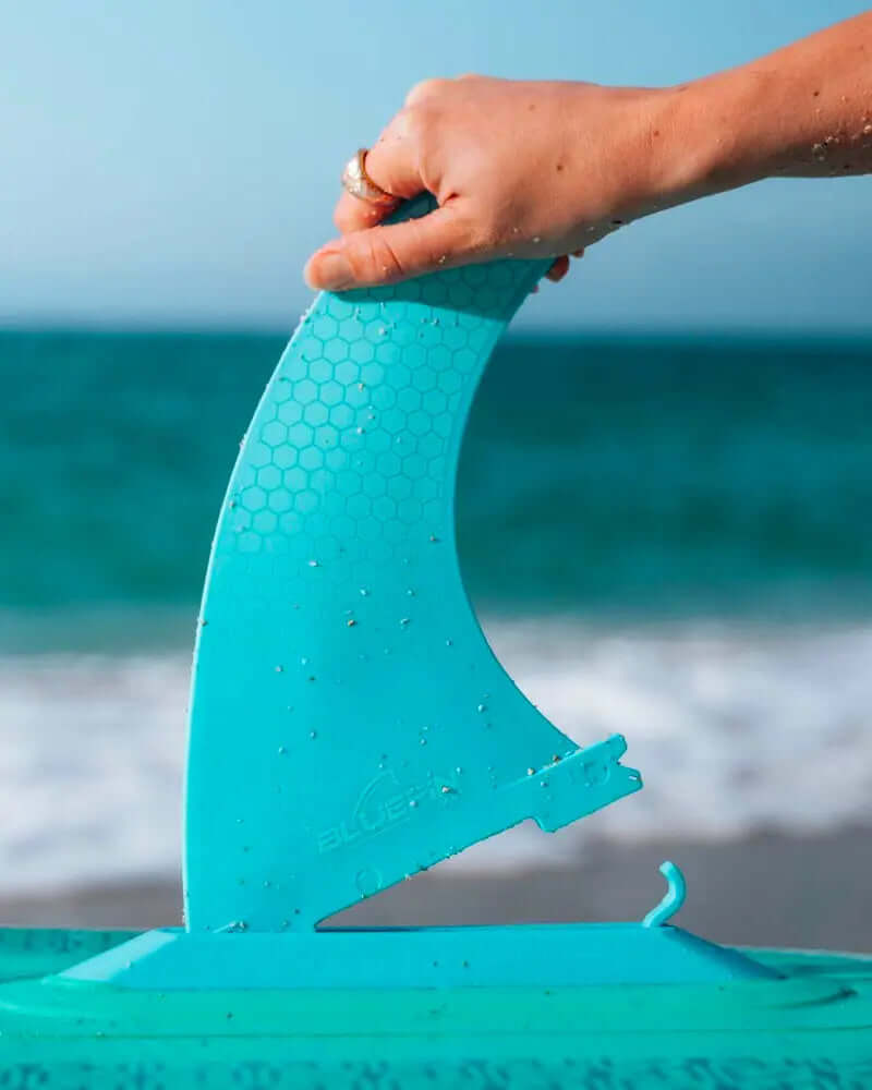 Hand holding a turquoise fin with a honeycomb texture above a paddleboard on the beach