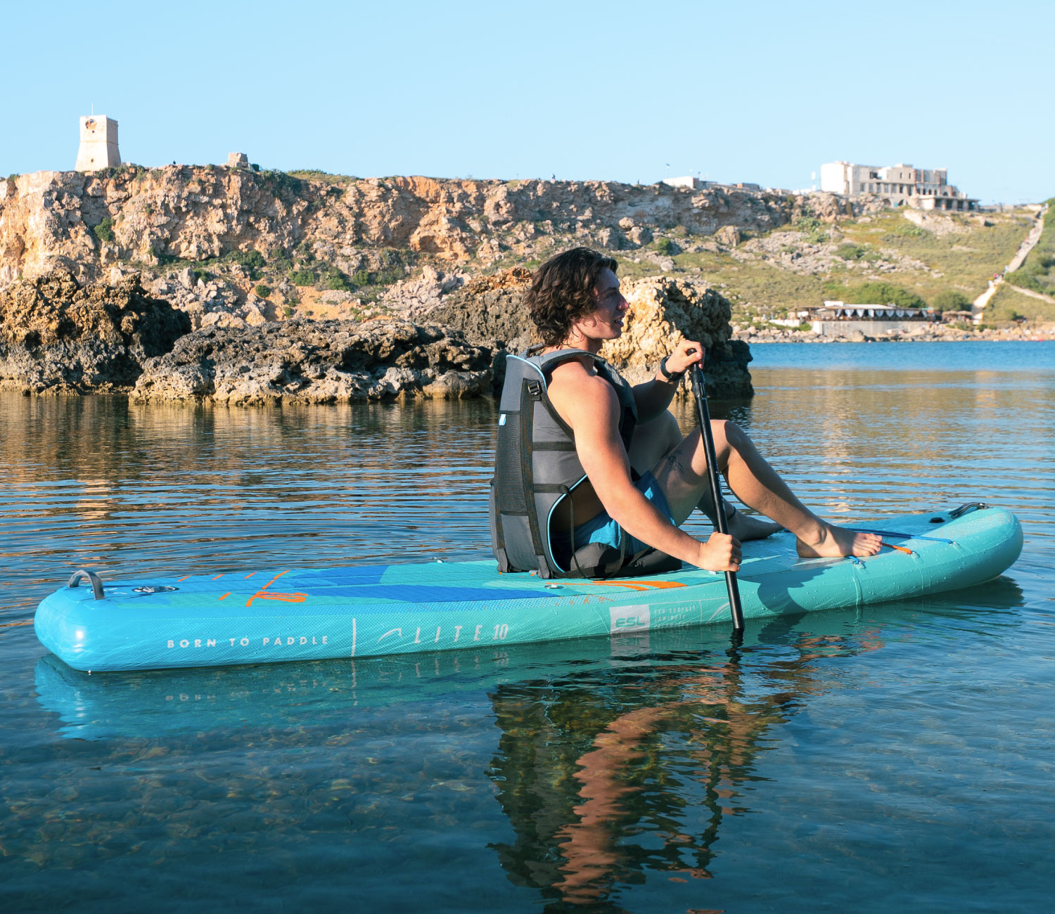 Inflatable turquoise paddleboard with a seated person paddling in calm water, rocky shore in background.