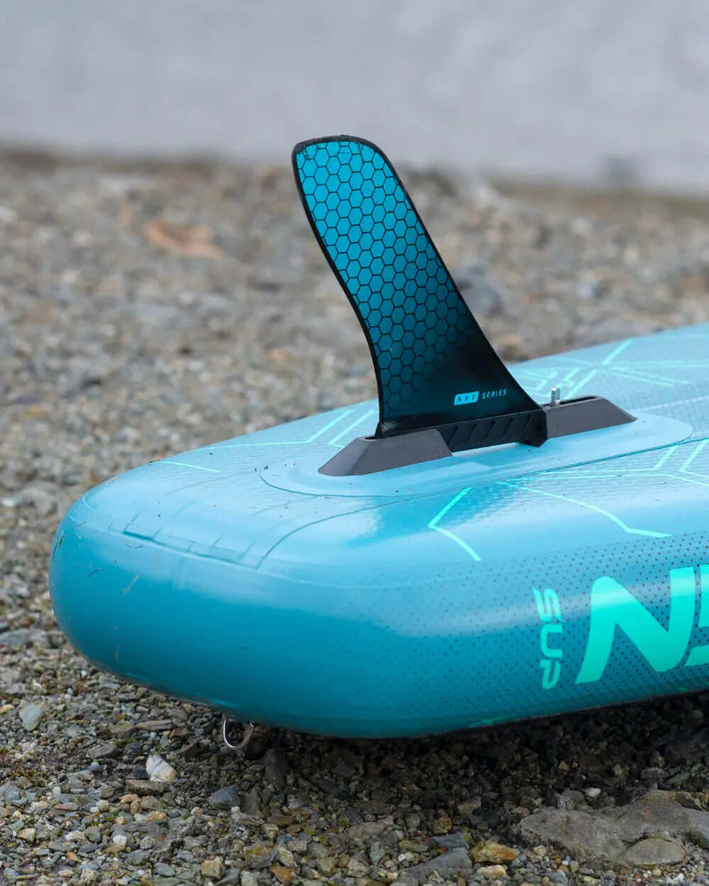 Inflatable paddleboard with a blue textured surface and a black center fin on a gravel background