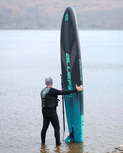 Person in wetsuit holding a Bluefin inflatable paddleboard in shallow water, wearing a life vest and beanie.