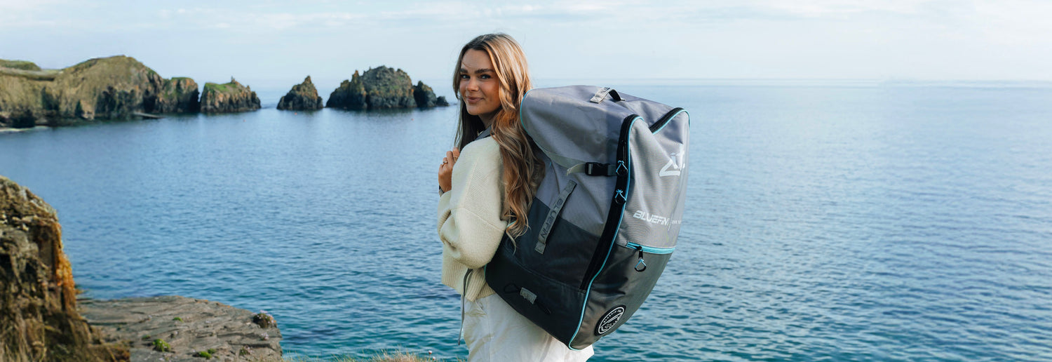 Woman wearing a white sweater, carrying a large gray Bluefin SUP backpack by the water.