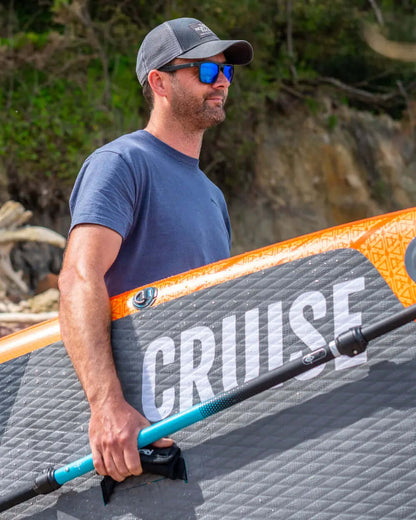 Man holding an inflatable paddleboard with orange edge and textured gray surface, wearing sunglasses and a cap.