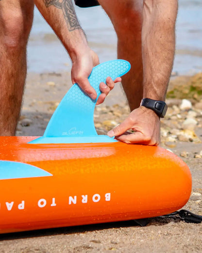 Person attaching a blue fin to an orange inflatable paddleboard on a sandy beach