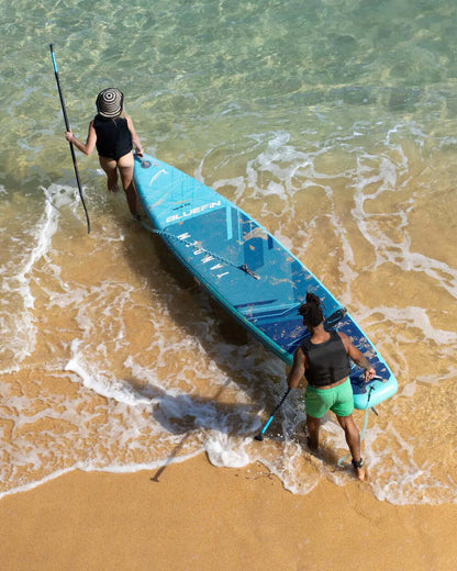 Two people wading in shallow water with a blue paddleboard and paddles, one wearing a hat.